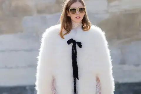 Estilo maximalismo: mujer con abrigo blanco de pelo, gafas grandes y lazo negro.