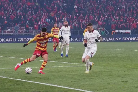 Piłkarz w żółto-czerwonych barwach strzela gola, podczas gdy zawodnik Jagiellonii Białystok w białej koszulce biegnie obok. Meczu Widzew Łódź – Jagiellonia Białystok.
