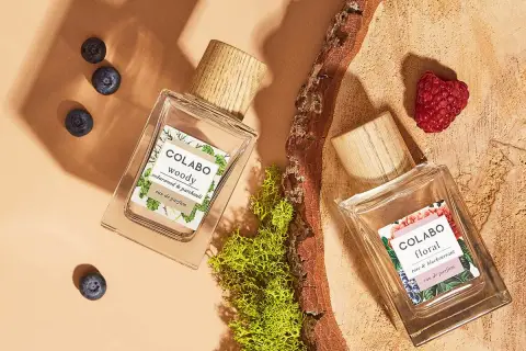 Dwa flakony perfum COLABO: "woody" z jagodami i "floral" z maliną, otoczone mchem i drewnem.