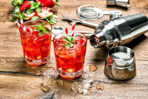 Dwa orzeźwiające drinki z martini rosato, lodem i truskawkami, obok shakera i akcesoriów barmańskich.