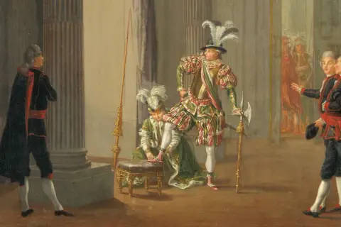 Scen från teater kungen där en person får sina skor på sig. Flera figurer i historiska dräkter.