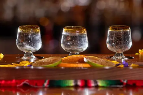 Trzy kieliszki z mezcalem, plasterki limonki i pomarańczy na drewnianej desce.