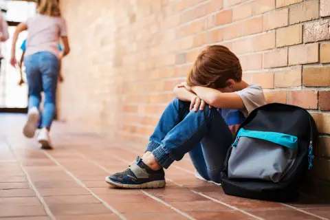 Chłopiec siedzi przy ceglanej ścianie, ukrywając twarz w dłoniach. W tle oddalają się inne dzieci. To obraz bullying po polsku.