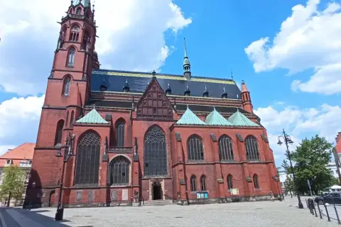 Gotycka katedra w mieście nad Kaczawą, z ceglaną fasadą i zielonymi dachami, na tle błękitnego nieba z chmurami.