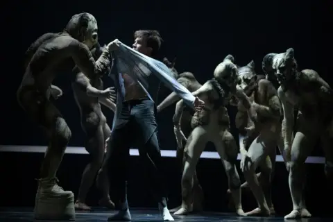Tancerz w Teatrze Wielkim w Warszawie, otoczony przez postaci w maskach, w scenie z repertuaru.