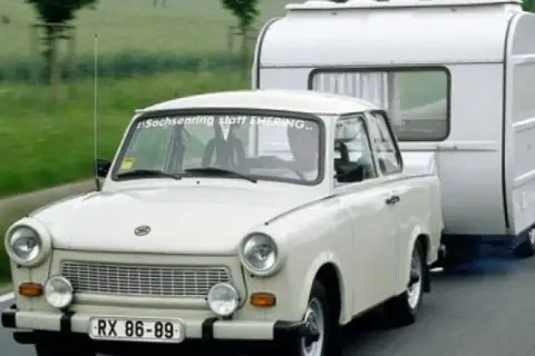 Biały trabant ciągnie przyczepę kempingową po drodze.