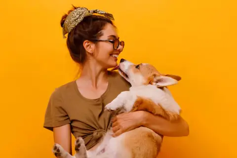 Uśmiechnięta dziewczyna z okularami i bandaną na głowie tuli corgi. Piesek liże ją po nosie. Wiele imion na N, jak Nela czy Nika, pasowałoby do tej słodkiej pary.