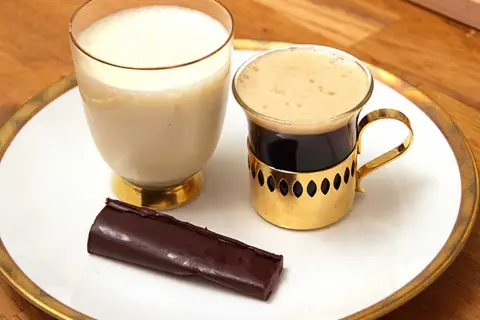 En bit mörk choklad ganache ligger bredvid en kopp kaffe och ett glas mjölk på en vit tallrik med guldkant.