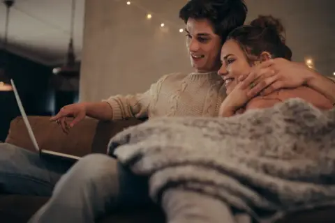Para młodych ogląda coś na laptopie, przytuleni pod kocem. Ich bliskość i uśmiechy sugerują, że historia o "fake dating" tak bardzo angażuje emocjonalnie czytelników, bo pozwala przeżywać rodzące się uczucia.