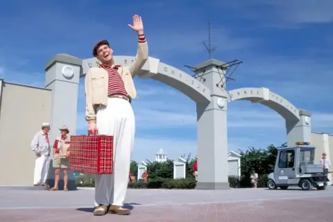 Truman Show obsada: Jim Carrey wita się z widzami, trzymając czerwoną walizkę. W tle łuk z napisem "Omnibus".