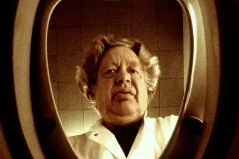 En man med lockigt hår, klädd i vit rock, tittar in i en toalettstol. Ernst Hugo Järegård i en minnesvärd scen.