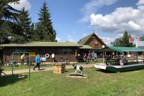 Na placu przed drewnianymi budynkami, wśród drzew, stoi łódź i krowa na moście. Je je jeleń witnica, jakby czekał na wycieczkę.