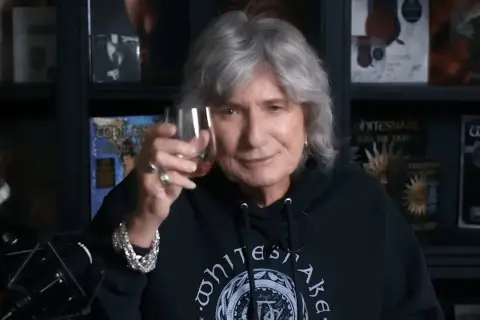 David Coverdale heute, mit grauem Haar und einem Glas in der Hand, trägt ein schwarzes Whitesnake-Hoodie.