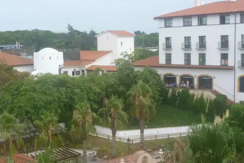 Edificio blanco con tejado naranja, palmeras y vegetación exuberante. Parece el hotel El Paso PortAventura, listo para recibir a sus huéspedes.