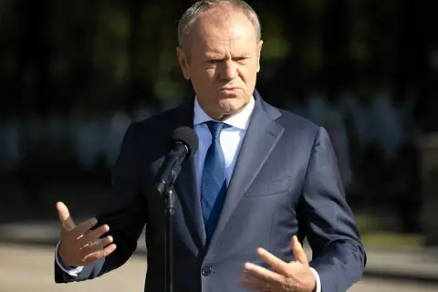 Donald Tusk w garniturze przemawia przy mikrofonie, gestykulując rękami.