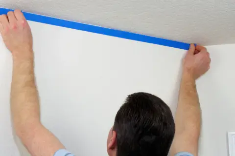 Mężczyzna przykleja niebieską taśmę malarską, by zamaskować krzywy sufit.