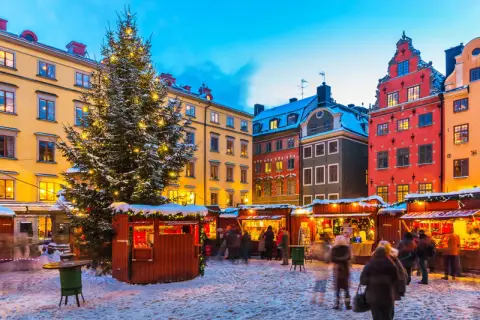 En snötäckt vinterviken julmarknad med ett stort, upplyst träd och färgglada hus. Folk strosar bland bodarna.