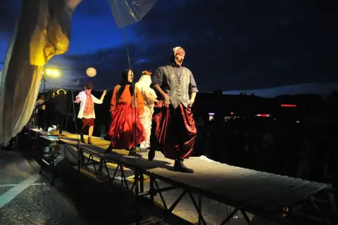 Aktorzy w barwnych strojach na scenie. W tle nocne niebo i widzowie. Spektakl "Teatr ósmego dnia" trwa.