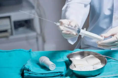 Lekarz przygotowuje się do amniopunkcji, trzymając strzykawkę z długą igłą. Na stole leżą też gaziki i inne akcesoria medyczne.