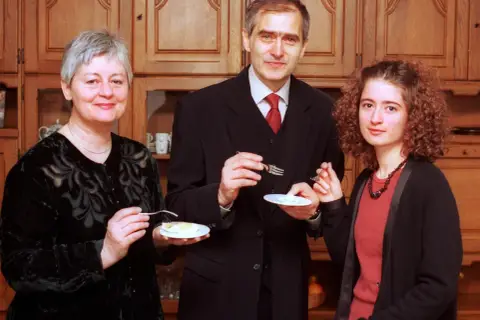 Trzy osoby: kobieta z krótkimi siwymi włosami, mężczyzna w garniturze i młoda kobieta z kręconymi włosami, jedzą coś z talerzyków.
