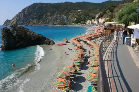 Plage animée à Moneglia, Italie, avec des parasols rayés, des baigneurs dans la mer turquoise et des collines verdoyantes en arrière-plan.