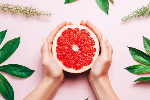 Dłonie trzymają połówkę grejpfruta, otoczone zielonymi liśćmi. Naturalne piękno i świeżość, jak wyciąg z pestek grejpfruta.