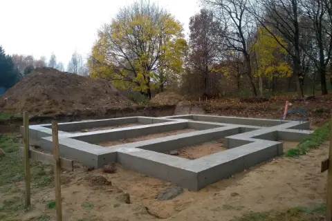 Fundament domu wylewany na ławach fundamentowych, poniżej strefa przemarzania gruntu. W tle jesienne drzewa i usypana ziemia.