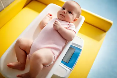 Noworodek w różowym body leży na wadze. Rodzice często zastanawiają się, ile powinien przybierać noworodek, by mieć pewność, że rozwija się prawidłowo.