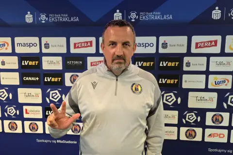 Trener Motoru Lublin z trzema palcami uniesionymi w geście zwycięstwa na tle sponsorów klubu.