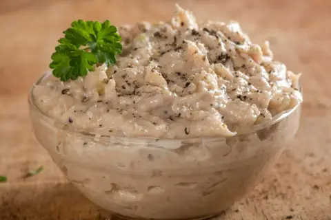 Pyszna pasta z tuńczyka, doprawiona pieprzem i udekorowana natką pietruszki, w szklanej miseczce na drewnianym tle.