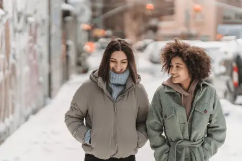 Deux amies discutent en marchant dans la neige. Une idée outfit hiver parfaite : doudoune beige et manteau vert ceinturé.