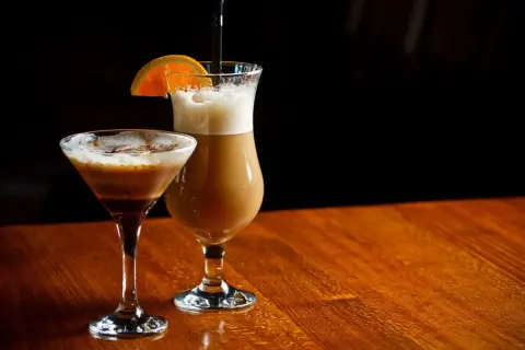 Dwa drinki na drewnianym stole: w kieliszku martini kawa z alkoholem i w wysokiej szklance latte z plasterkiem pomarańczy.