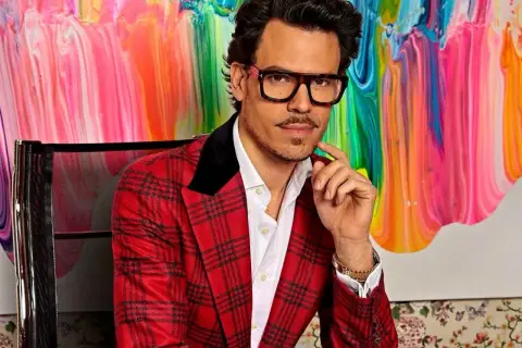 Juan Avellaneda, con su característico traje a cuadros rojos y gafas llamativas, posa con una mano en la barbilla frente a un vibrante cuadro abstracto.
