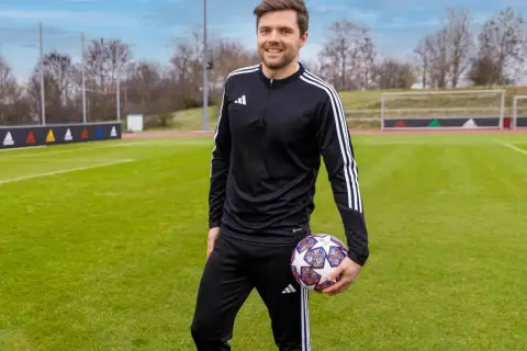 Uśmiechnięty trener piłki nożnej w czarnym dresie z białymi paskami, trzymający piłkę na zielonym boisku.
