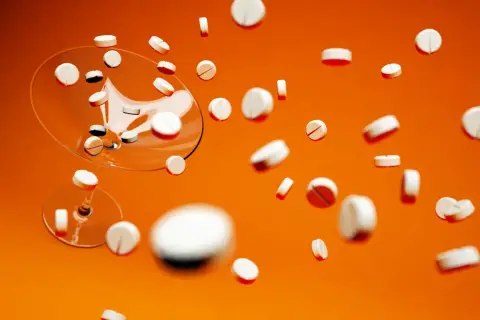 Białe tabletki, w tym Xarelto 20 mg, spadają z kieliszka na pomarańczowym tle.