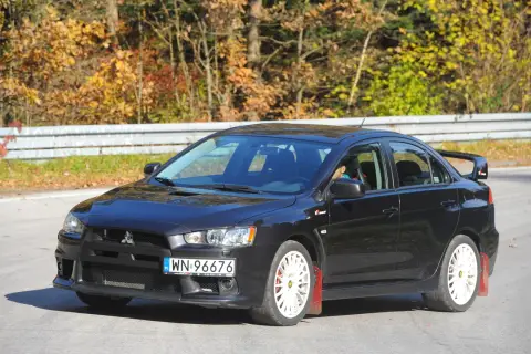 Czarny Mitsubishi Lancer 9 z białymi felgami i czerwonymi chlapacami na tle jesiennego lasu.