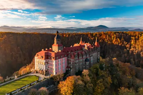 Malowniczy hotel zamek otoczony jesiennym lasem, z wieżą i czerwonymi dachami, na tle gór i błękitnego nieba.