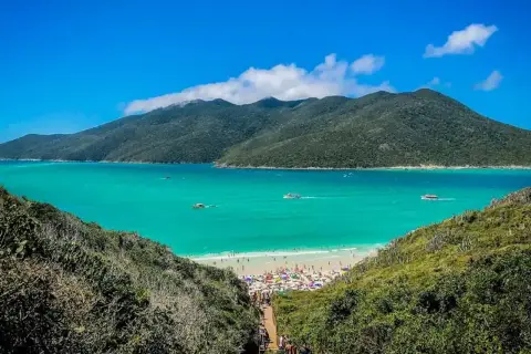 Escenario idílico para viajar a Brasil: playa con aguas turquesas, montañas verdes y gente disfrutando bajo sombrillas.