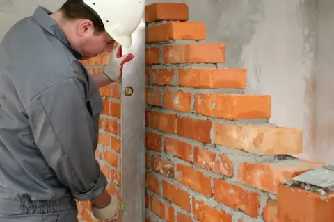 Murarz w kasku i rękawicach sprawdza pion muru. Cegły o standardowych wymiarach układane są na zaprawie.