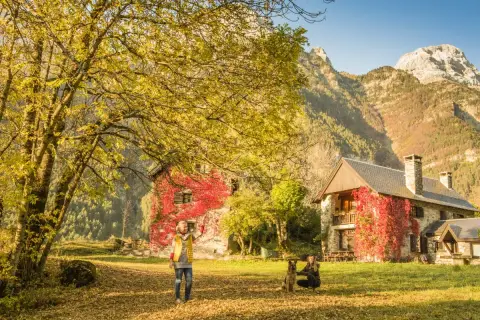 Dos mujeres y un perro disfrutan del otoño en el valle de Pineta, ideal para rutas. Las casas cubiertas de hiedra roja y las montañas crean un paisaje idílico.