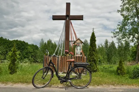 Rower przy kapliczce w Hajnówce, Białowieża. Drewniany krzyż z figurką Chrystusa i Matki Boskiej, otoczony zielenią i polnymi kwiatami.