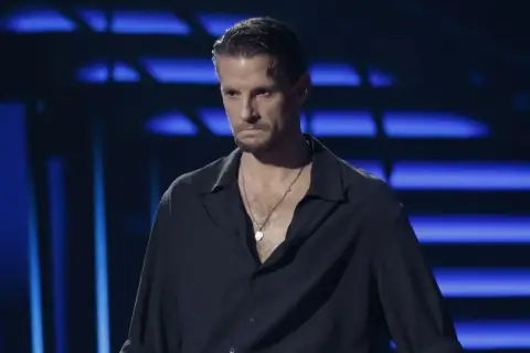 Sebastian Fabijański w czarnej koszuli z naszyjnikiem na tle niebieskich świateł.