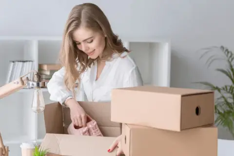 Kobieta rozpakowuje paczkę InPost, wyjmując z niej różowe buty. Obok stos kartonów.