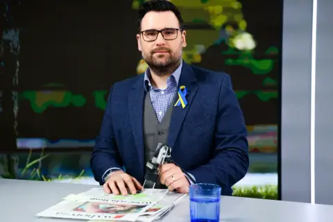 Michał Szajder, dziennikarz, w garniturze z przypinką w barwach Ukrainy, siedzi przy stole z gazetami i szklanką wody.