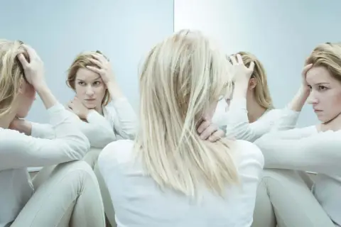 Kobieta z blond włosami w białej bluzce, trzymająca się za głowę, otoczona swoimi odbiciami w lustrze.