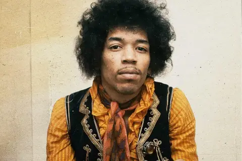 Jimi Hendrix w pomarańczowej koszuli i kamizelce, z charakterystyczną fryzurą. W tle subtelna, lekko zatarte tło, jakby w **purple haze**.