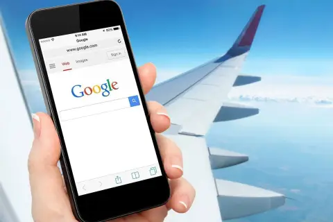 Dłoń trzyma smartfon z otwartą wyszukiwarką Google, na tle skrzydła samolotu i nieba. Czy w samolocie można mieć internet? Tak, można!