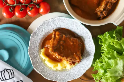 Pyszny sos z żeberek podany z purée ziemniaczanym na ozdobnym talerzu. Obok świeża sałata i pomidory.
