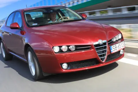 Czerwone Alfa Romeo 159 1.9 JTD pędzi po drodze, kierowca skupiony na trasie.
