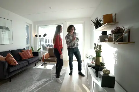 Två personer diskuterar olika inredningsstilar i ett ljust vardagsrum med modern möblering, en mörk soffa och hyllor med växter.